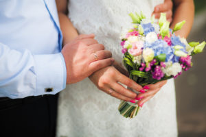 Wedding bouquet of flowers in hands of beautiful anonymous young bride. Wedding decor. Groom and bride enjoying each other. Just married happy couple hugging outside on summer or spring warm sunny day