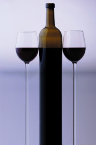 Stretched bottle and two glass goblets with red grape wine standing close to each other in studio isolated on white and grey backgroung, vertical picture